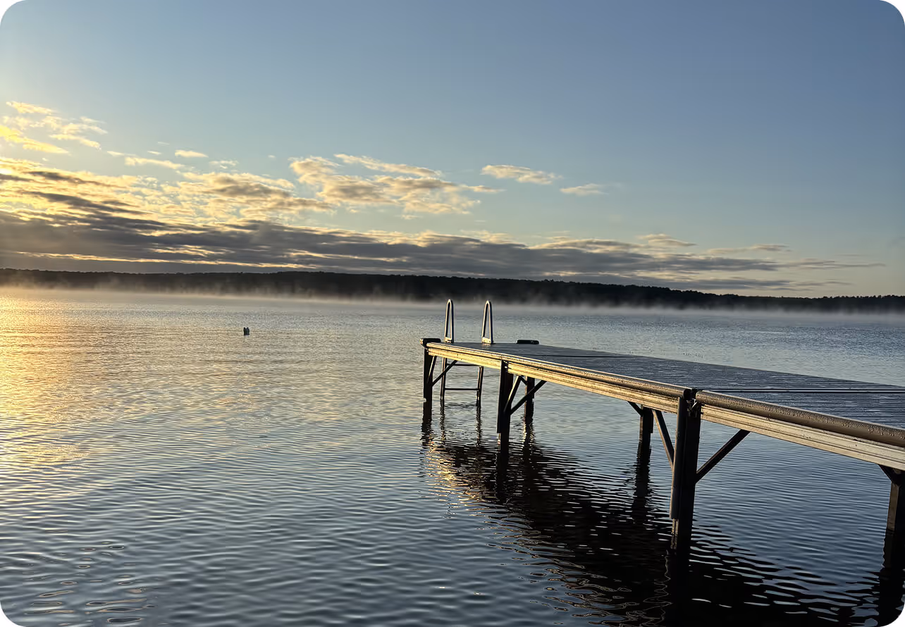 Wooden dock extending over calm lake water at sunrise with mist rising near tree-lined horizon.