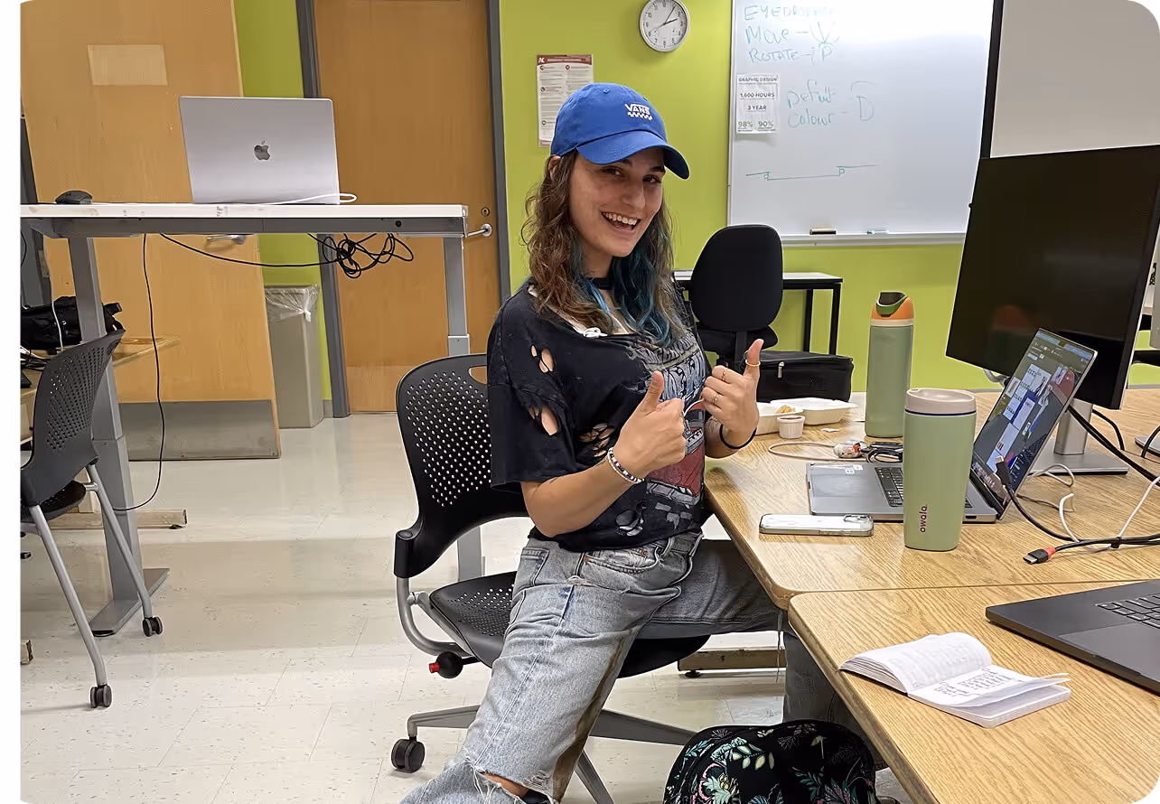 Person wearing a blue cap and ripped black t-shirt sitting on a chair at a desk giving two thumbs up in a classroom.