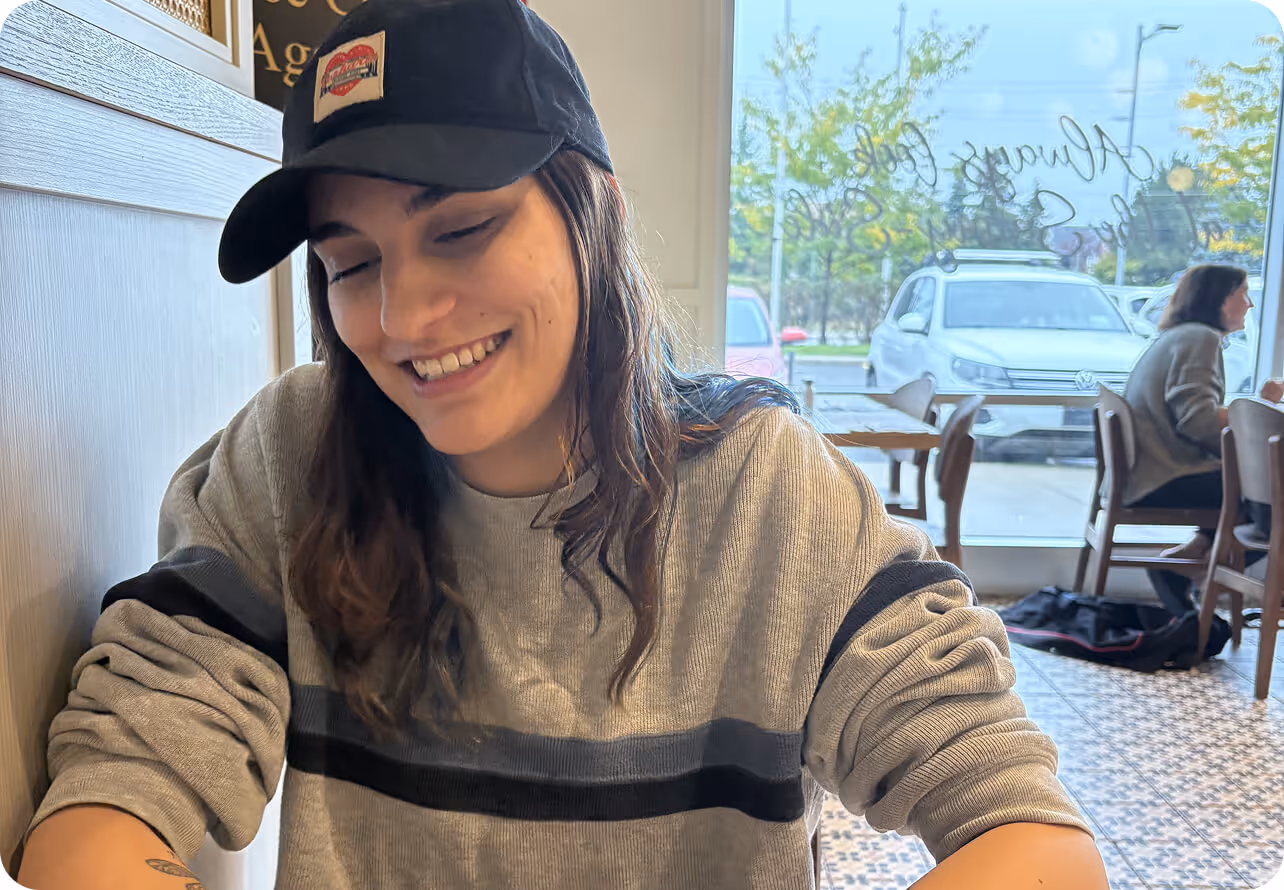 Smiling young woman wearing a black cap and gray sweater sitting inside a cafe near a window with parked cars visible outside.