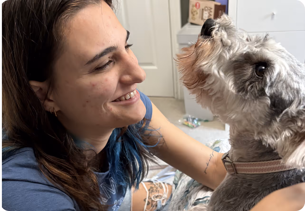 Smiling young woman with long dark hair and blue highlights looking at a gray and white dog wearing a collar indoors.