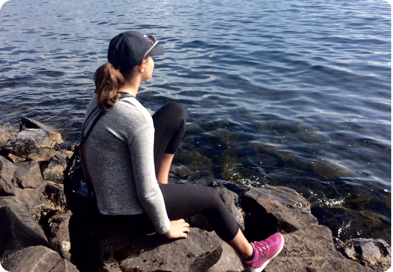 Woman in a gray top, black leggings, and purple sneakers sitting on rocks by calm water, wearing a black cap and sunglasses on her head.