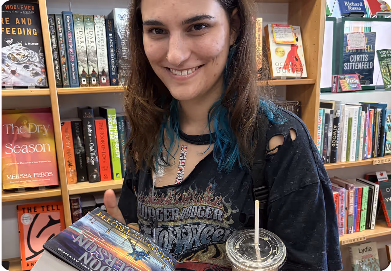 Smiling young woman with blue highlights holding books and a drink, standing in front of a bookstore shelf.