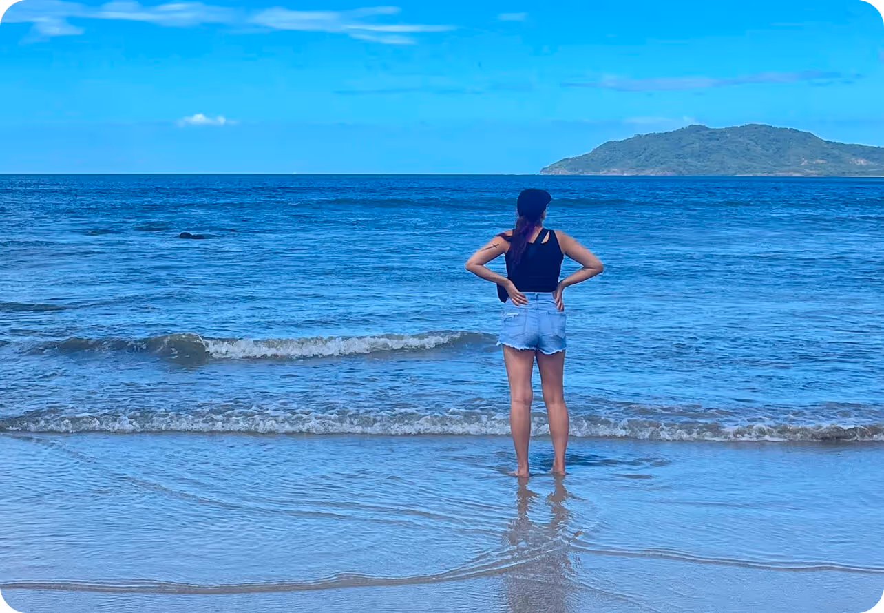 Person standing barefoot at the edge of the ocean, facing a calm sea with a distant island under a clear blue sky.