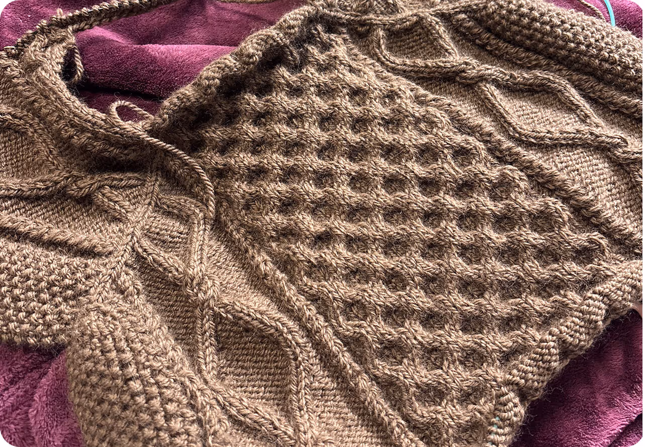 Close-up of a brown knitted garment with intricate cable and honeycomb patterns lying on a purple fabric.