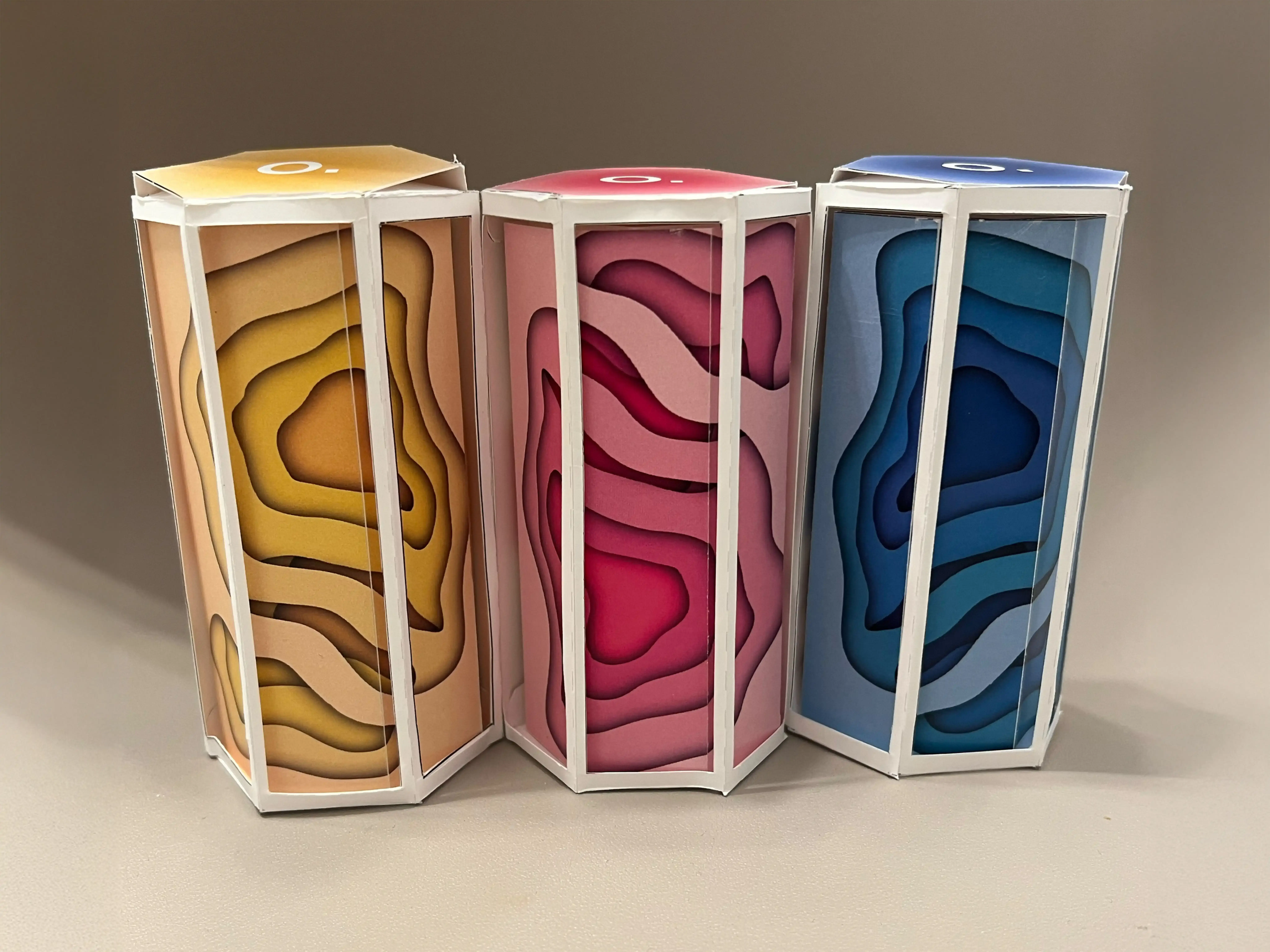 Three hexagonal prism packages with layered paper designs in yellow, pink, and blue, each with a white frame and a letter 'O.' on top.