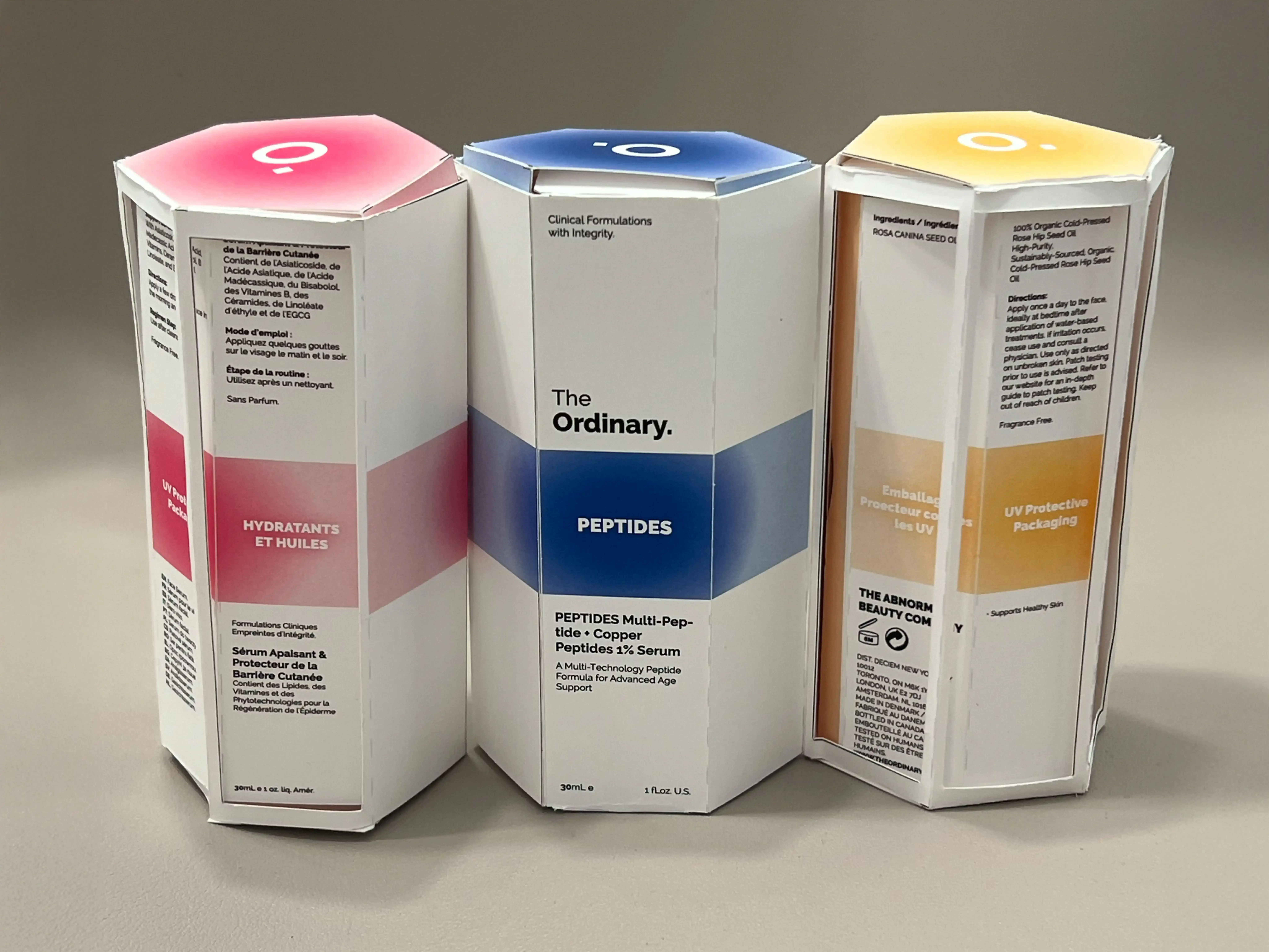 Three hexagonal skincare product boxes from The Ordinary, labeled Hydratants et Huiles, Peptides, and UV Protective Packaging, standing side by side.