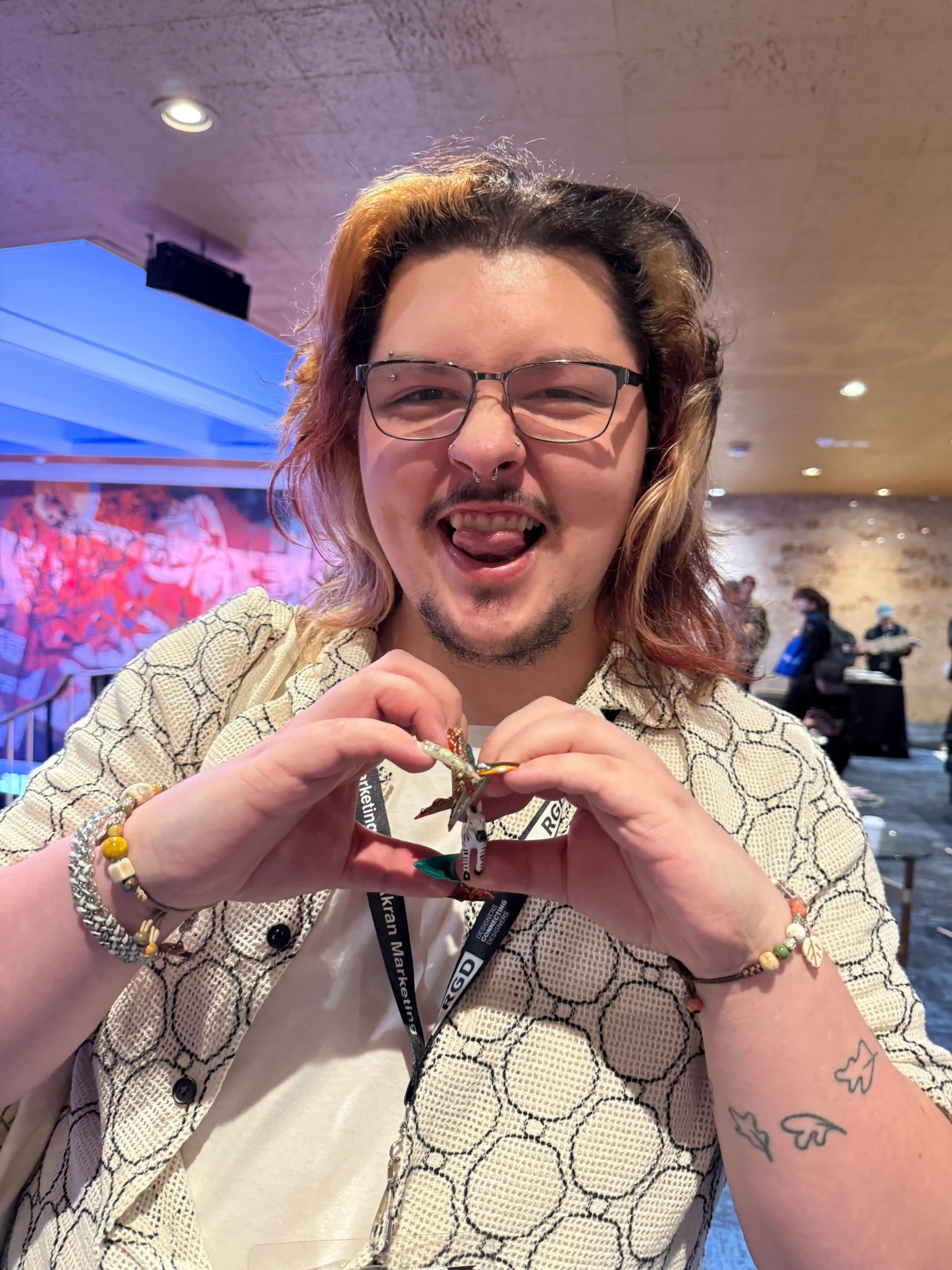 Alexander Bridson with glasses and dyed hair making a heart shape with hands around a small zebra figurine, smiling with tongue out indoors.