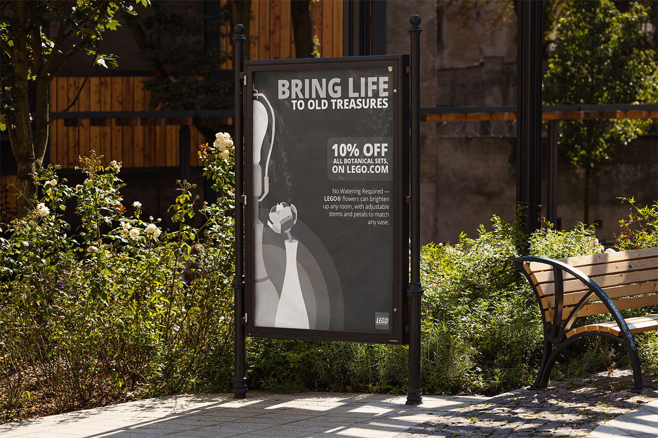 Outdoor black-framed sign promoting LEGO botanical sets with 10% off, next to a wooden bench and plants.
