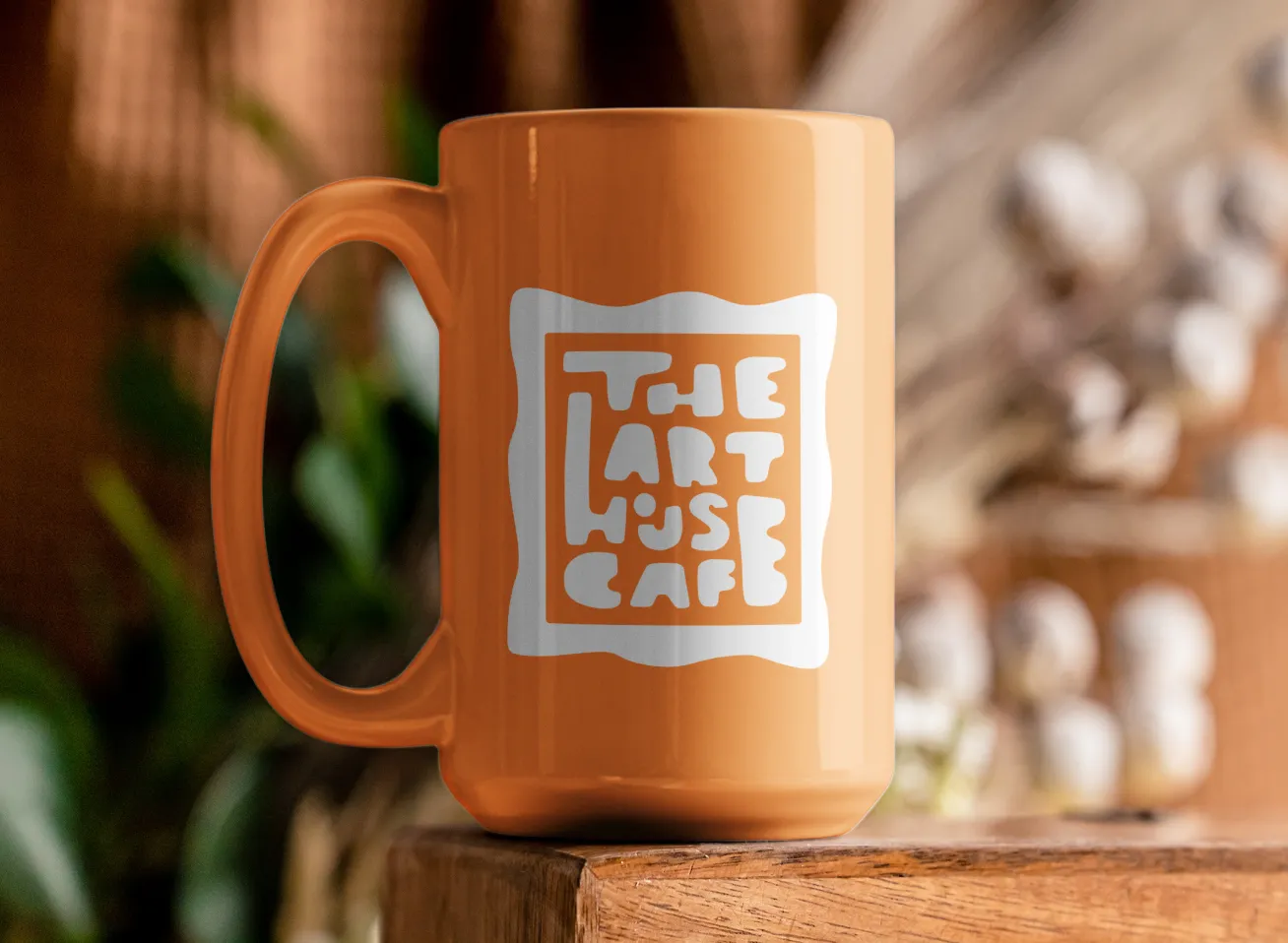 Orange ceramic mug with a white logo reading 'The Art House Cafe' sits on a wooden surface with blurred indoor plants and decor in the background.