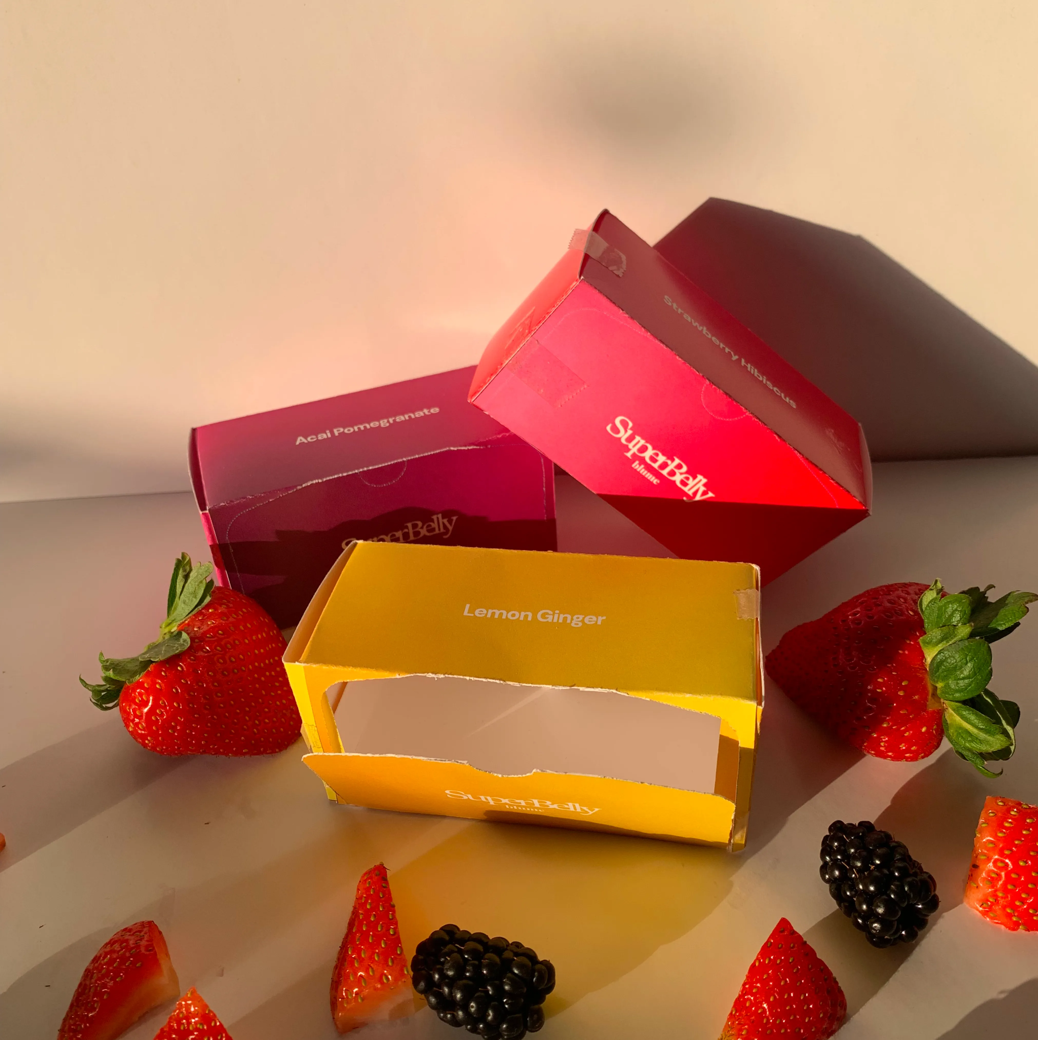 Three colorful SuperBelly product boxes labeled Acai Pomegranate, Strawberry Hibiscus, and Lemon Ginger surrounded by fresh strawberries and blackberries.