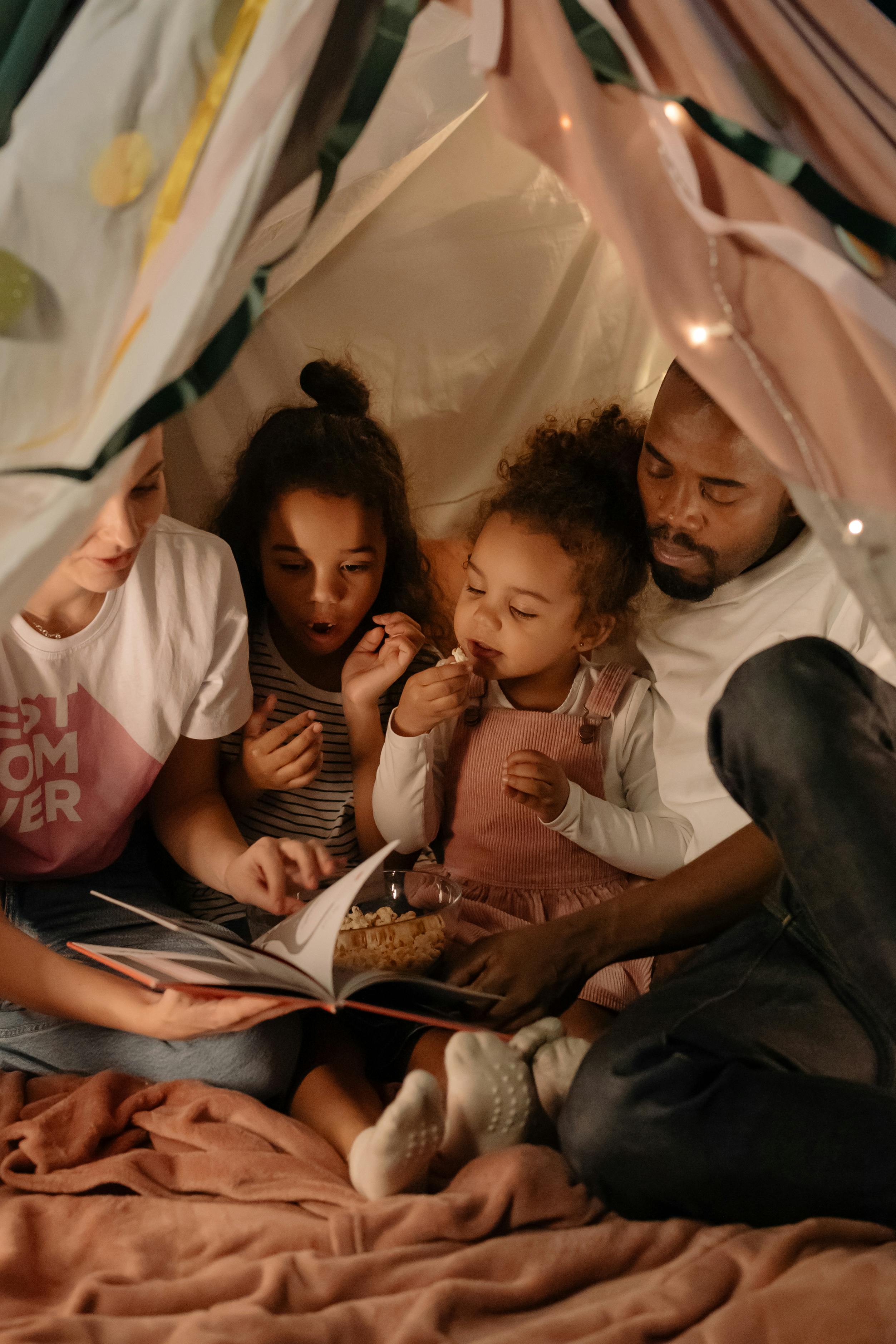 A family of four sitting inside a fabric tent, reading a book and eating popcorn together.