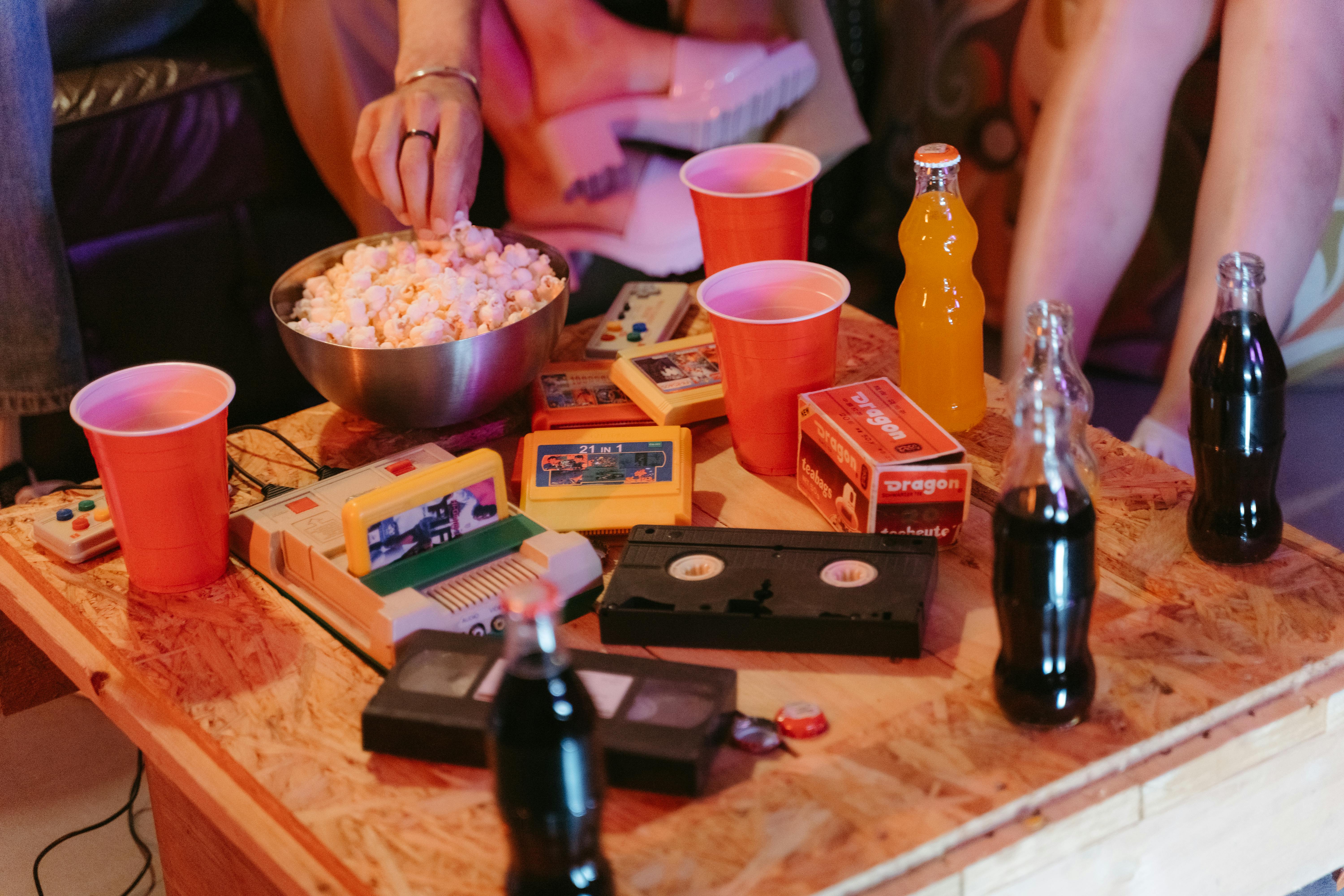 Wooden table with retro gaming cartridges, video game console, popcorn bowl, cassette tape, red cups, and soda bottles with people sitting around.
