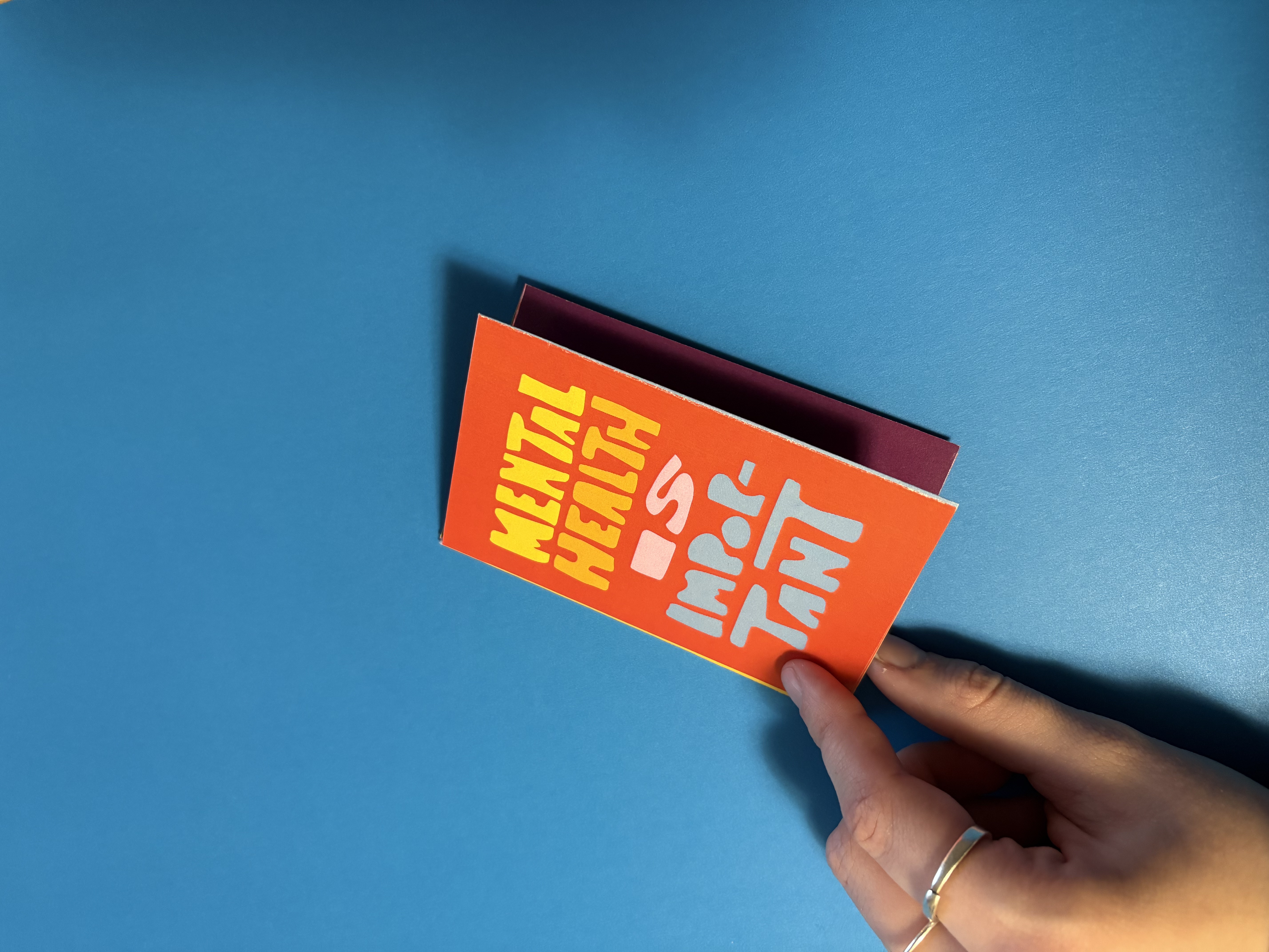 Hand holding a small orange booklet with yellow and white text reading 'Mental Health is Important' against a blue background.