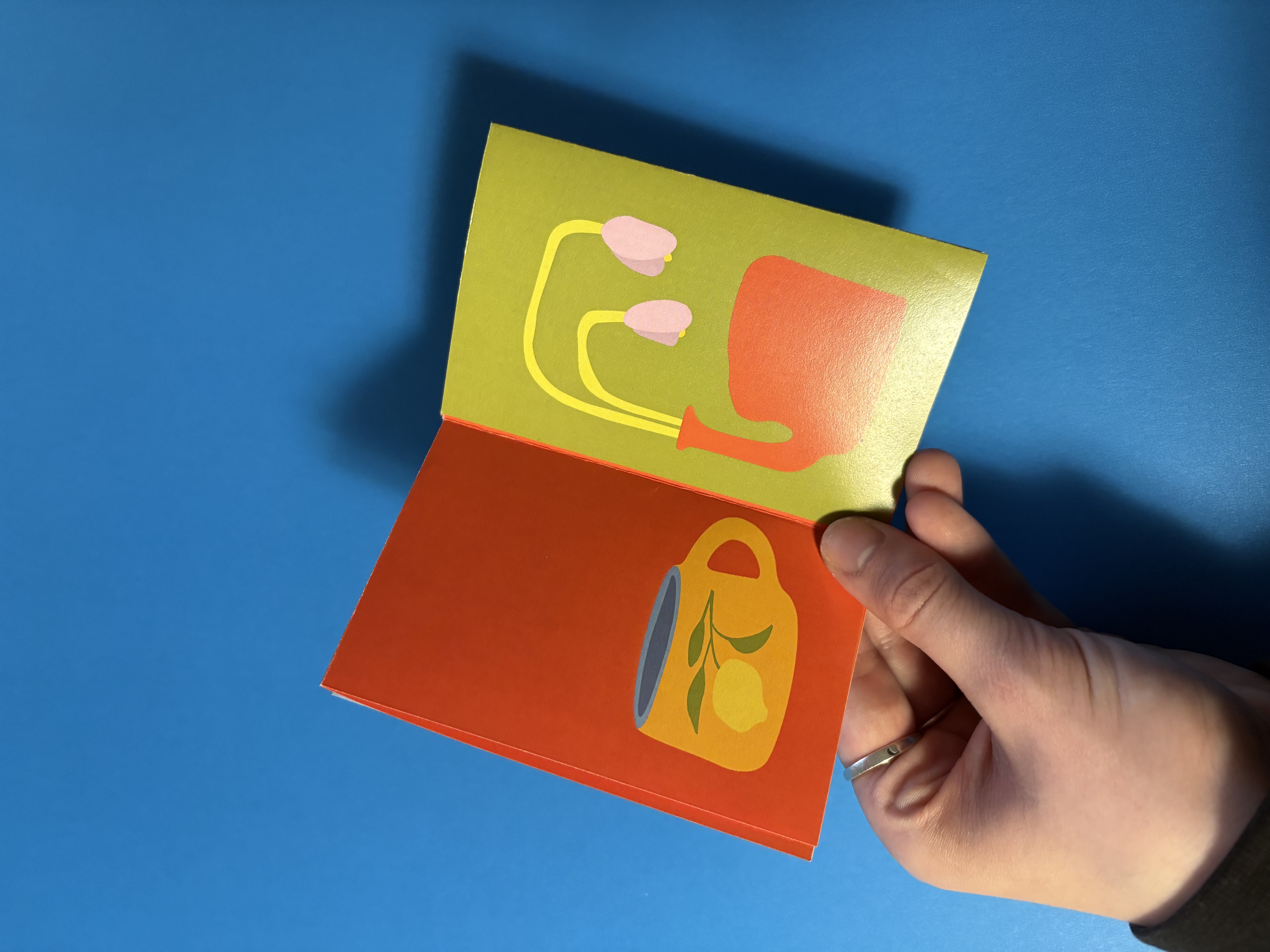 Hand holding an open card with a yellow cup and green leaves illustration on the left and a red watering can with drooping pink flowers on the right against a blue background.
