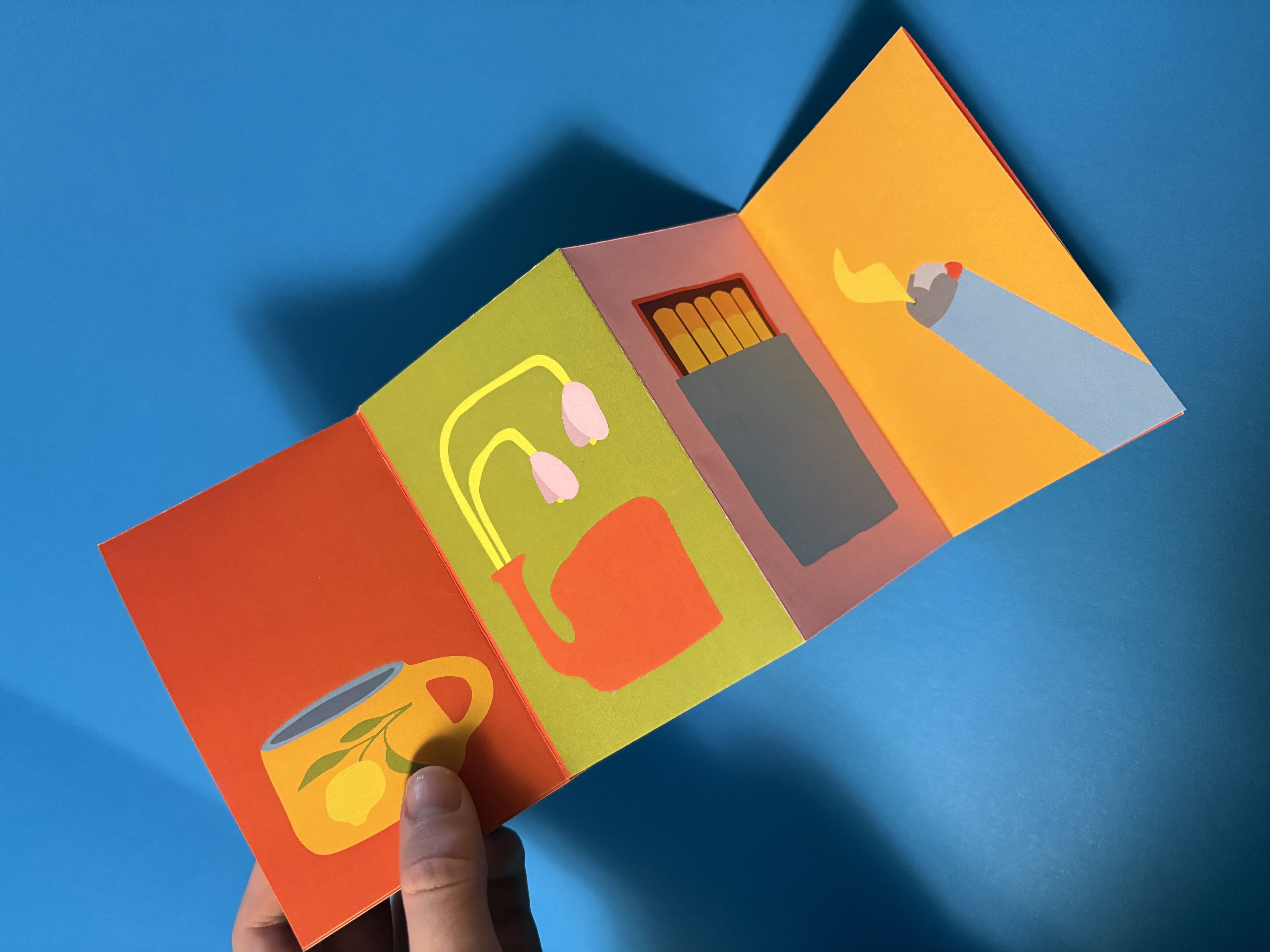 Hand holding a colorful folded card with illustrations of a yellow flowered mug, a red teapot with pink flowers, a matchbox with matches, and a lit cigarette on a blue background.