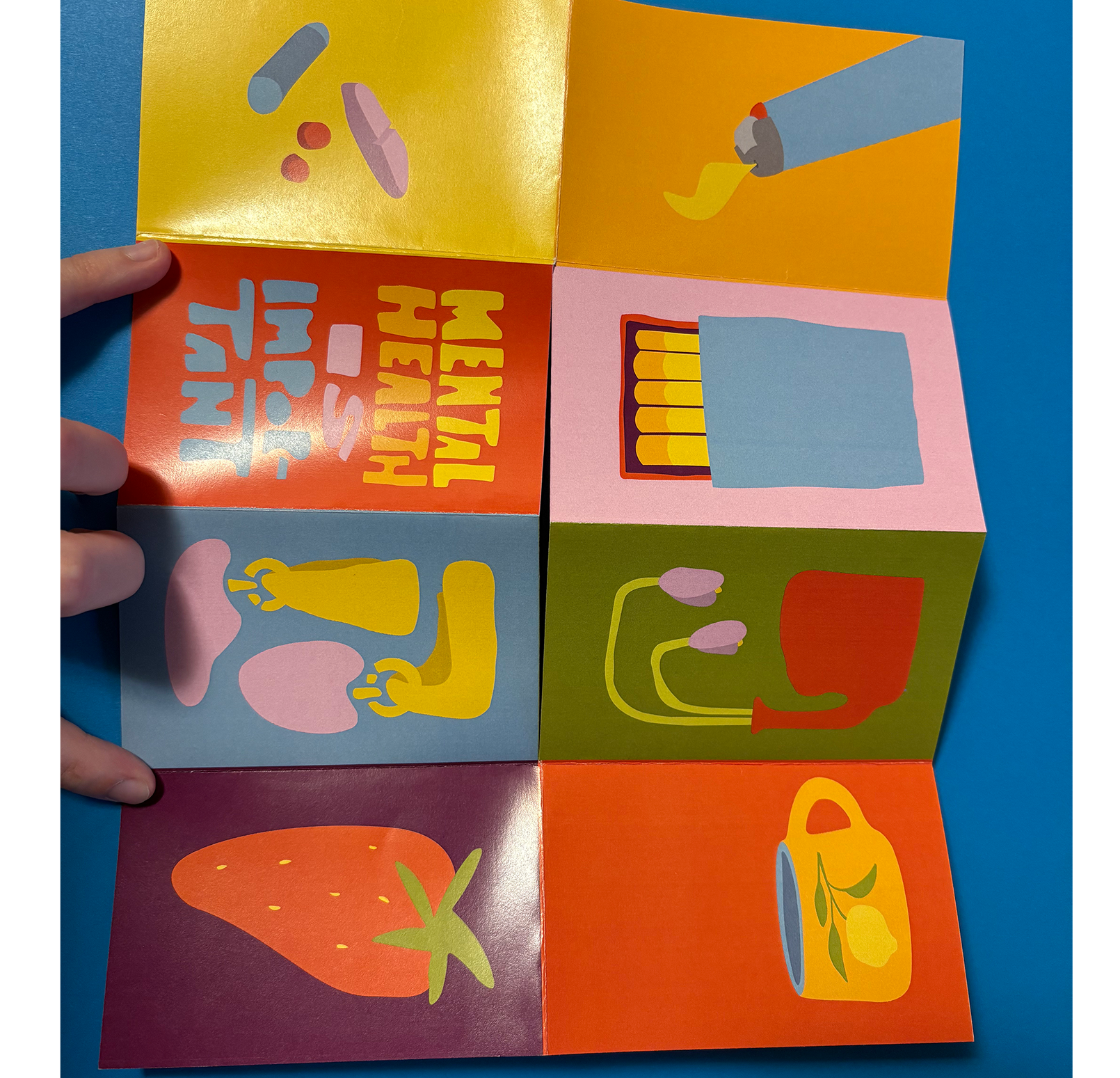 A colorful folded paper art piece featuring illustrations of pills, a paint tube, matchsticks, flowers, a red pipe, a strawberry, a mug with a lemon design, and text reading 'MENTAL HEALTH IS TINY'.