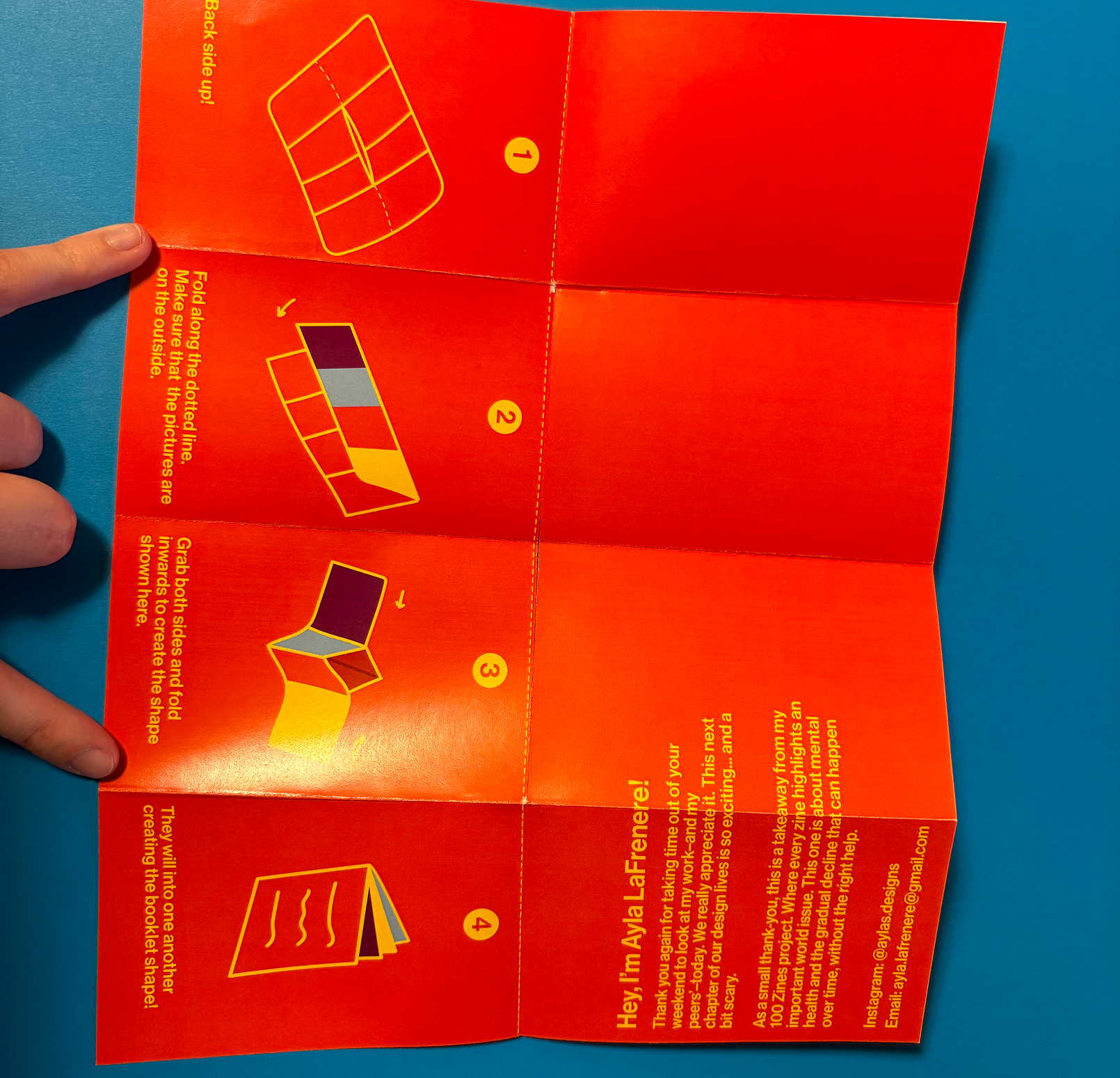Folded orange instruction sheet on blue surface showing four illustrated steps to fold a paper booklet, with a person's fingers holding the left edge.