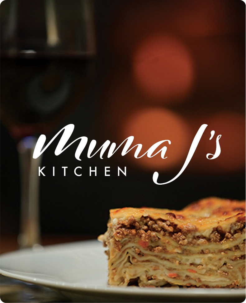 Slice of lasagna on a white plate with a blurred glass of red wine and warm bokeh lights in the background, overlaid with the text 'Muma J's Kitchen'.