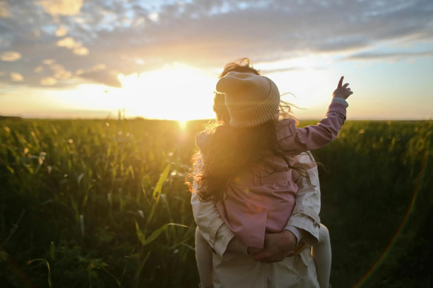 Person holding a child wearing a yellow knit hat and pointing toward the sunset over a green field.