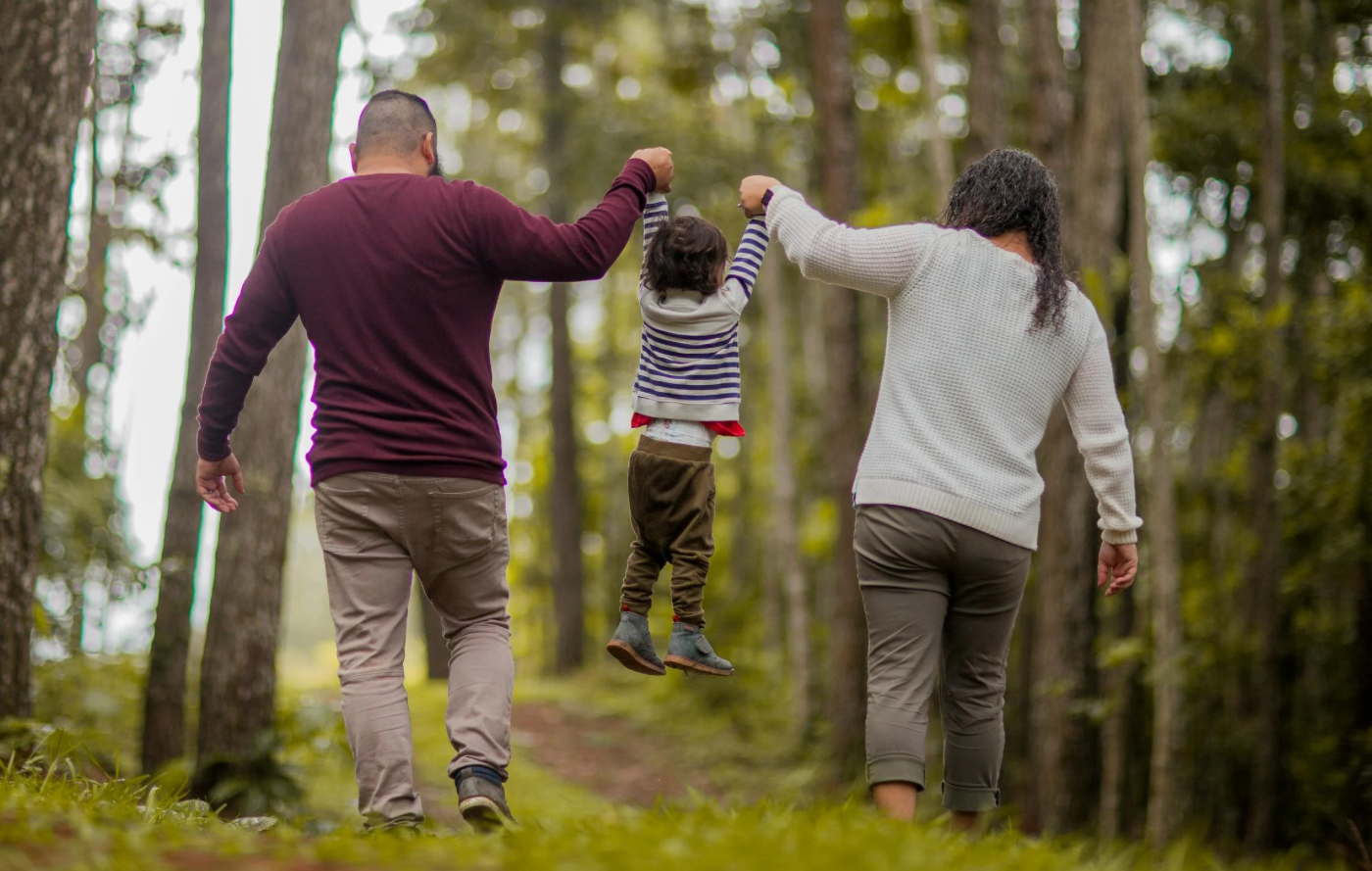 A couple walks through a forest holding a young child by both hands, lifting the child off the ground.