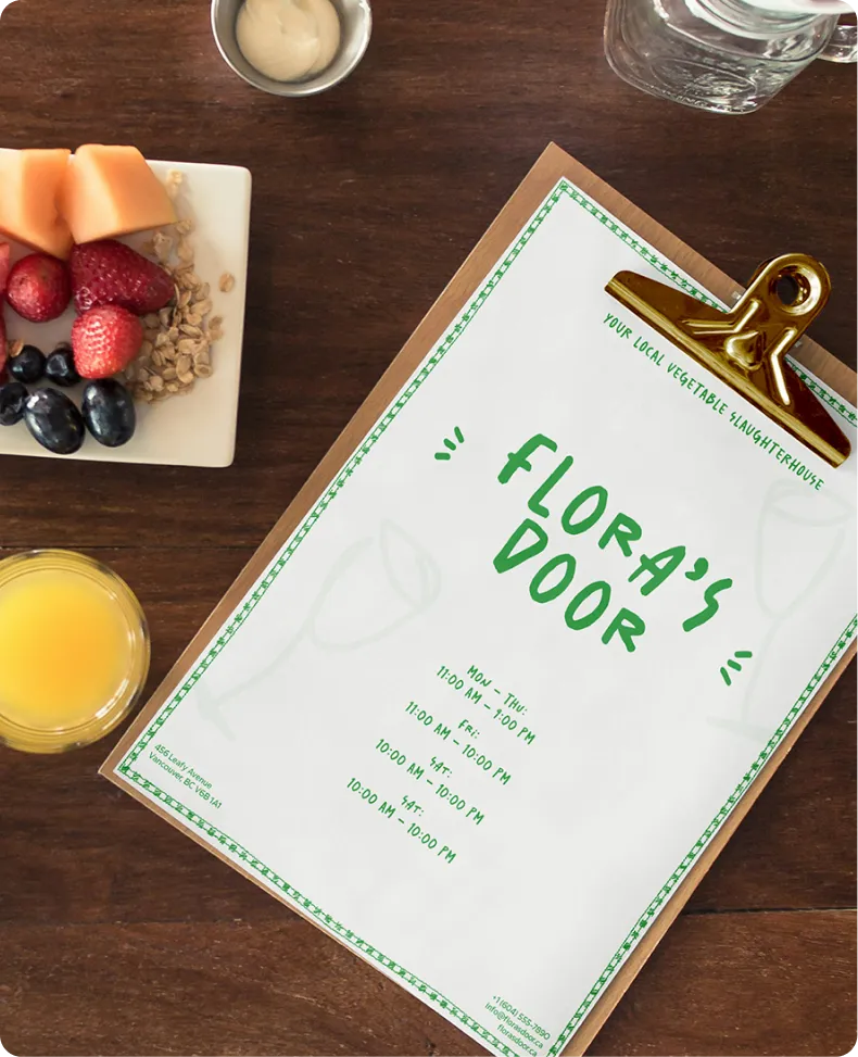 Clipboard menu for Flora's Door restaurant on a wooden table, accompanied by a plate of fresh fruit, a glass of orange juice, and a small dish of sauce.