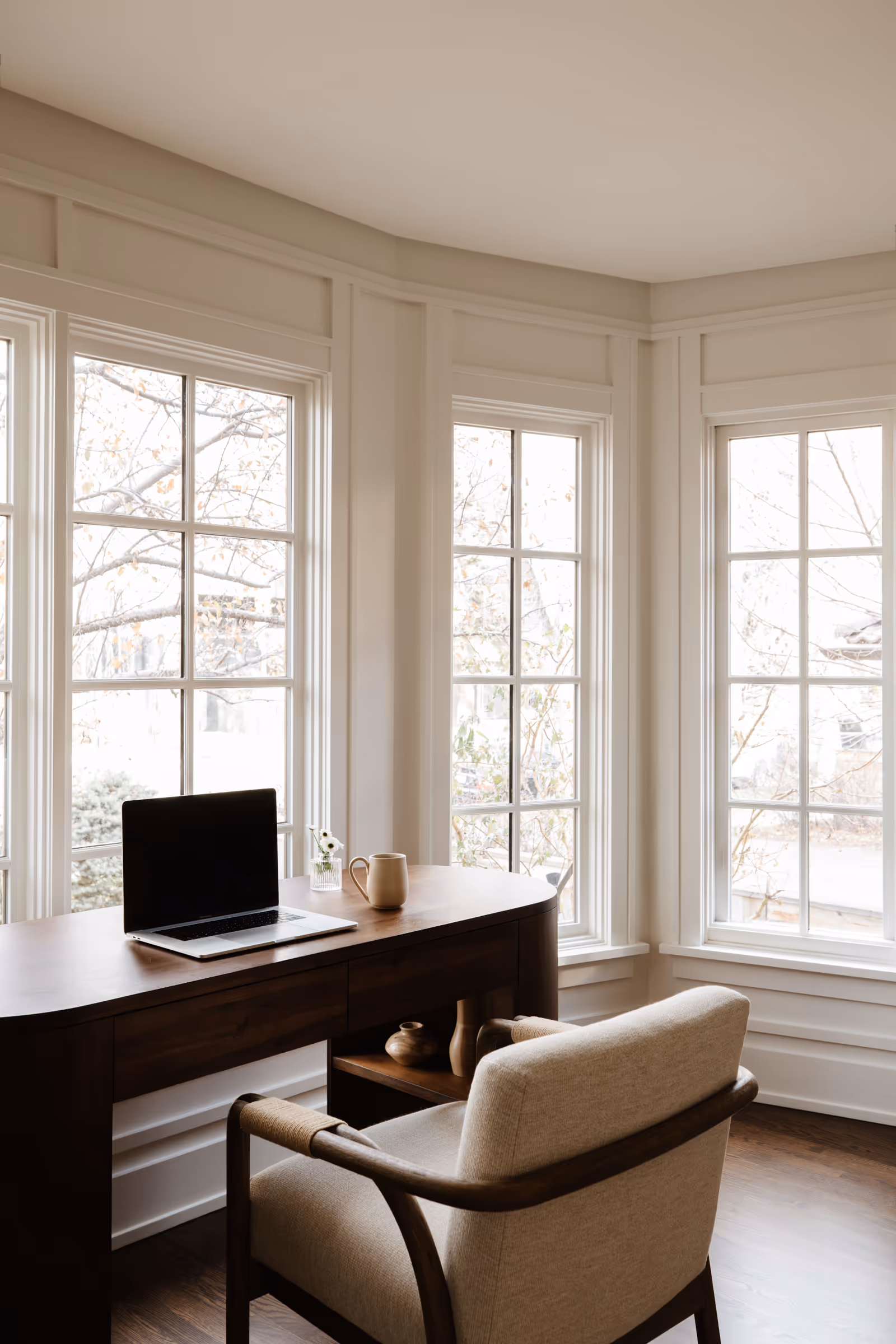 Modern home office with wooden desk, beige upholstered chair, laptop, mug, and large windows showing bare tree branches outside.