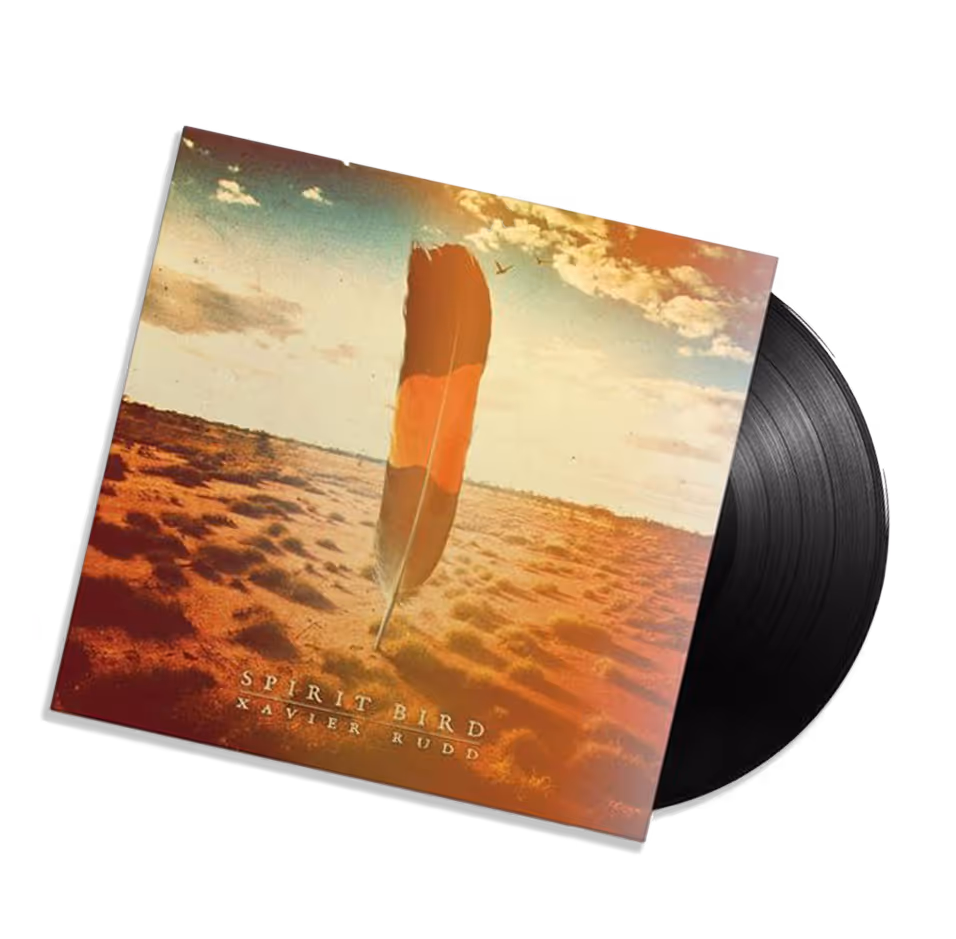 Vinyl record partially out of the cover featuring a large feather standing upright in a desert landscape with a blue sky and clouds, titled Spirit Bird by Xavier Rudd.