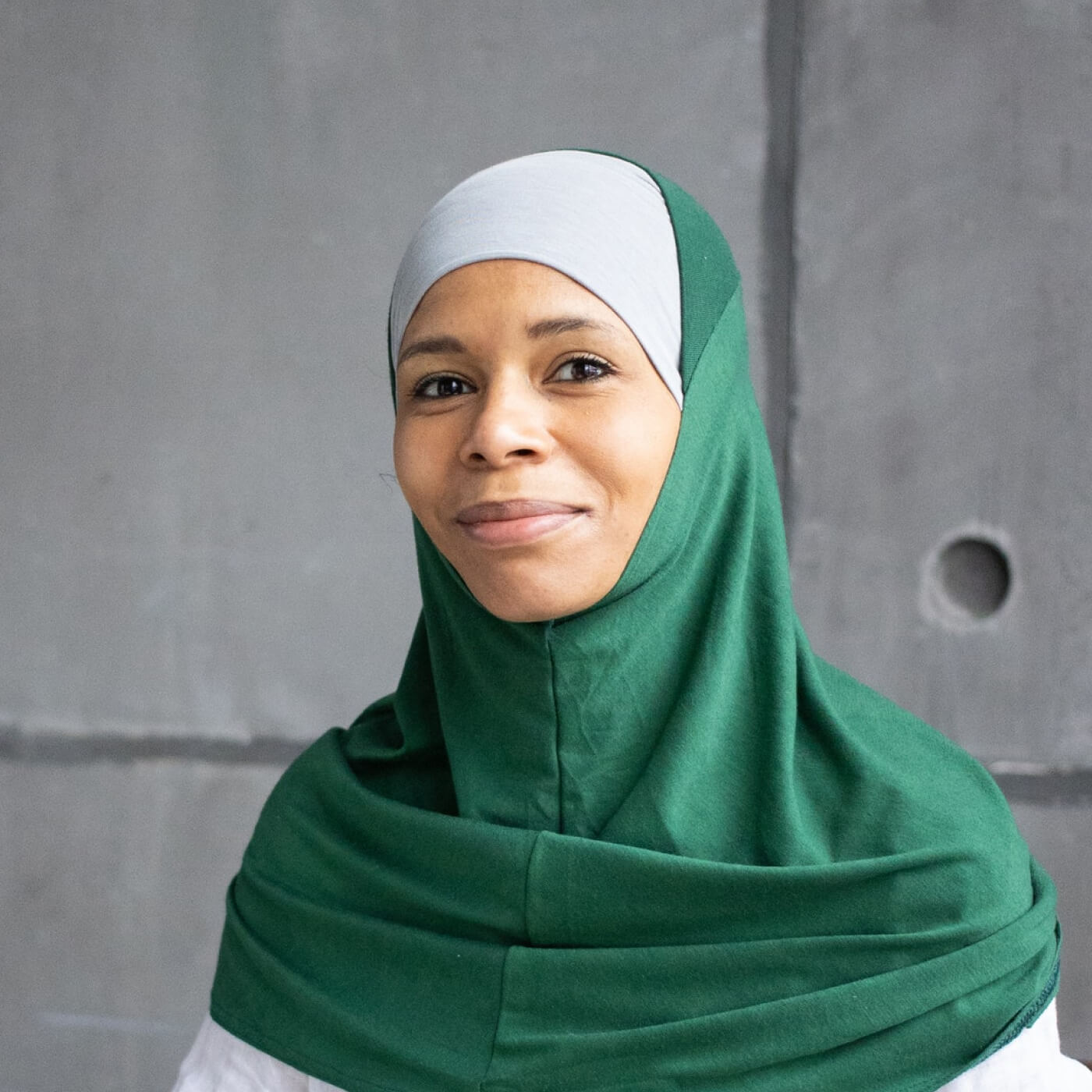 Portrait of a woman wearing a green hijab and light gray under-scarf against a gray concrete wall.