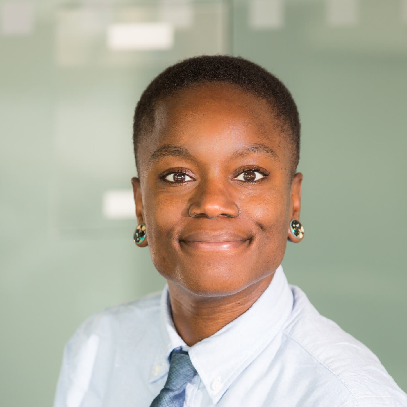 Smiling person with short hair wearing a light blue shirt and tie against a plain background.