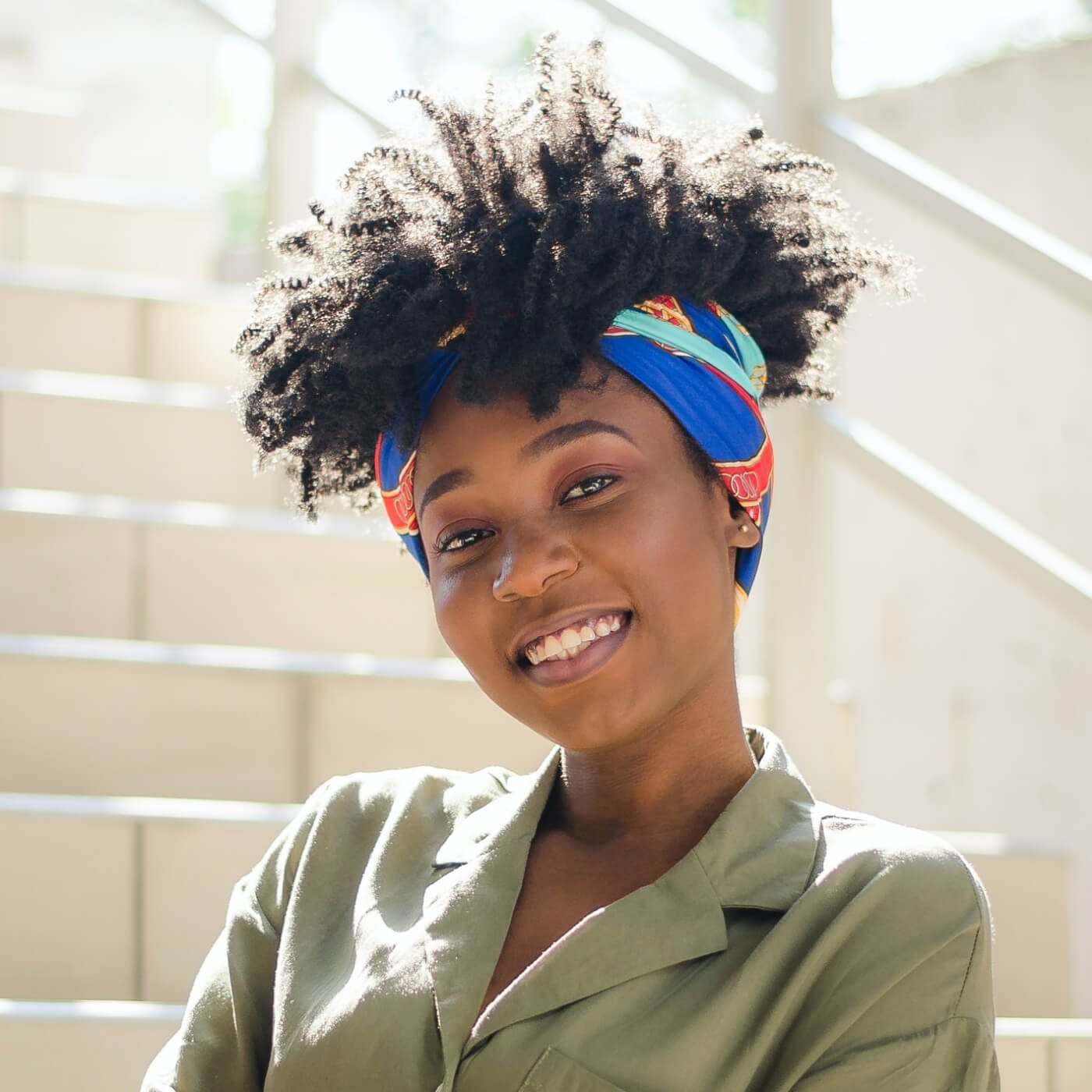 Smiling woman with natural curly hair wearing a colorful headband and olive green shirt, standing outdoors.