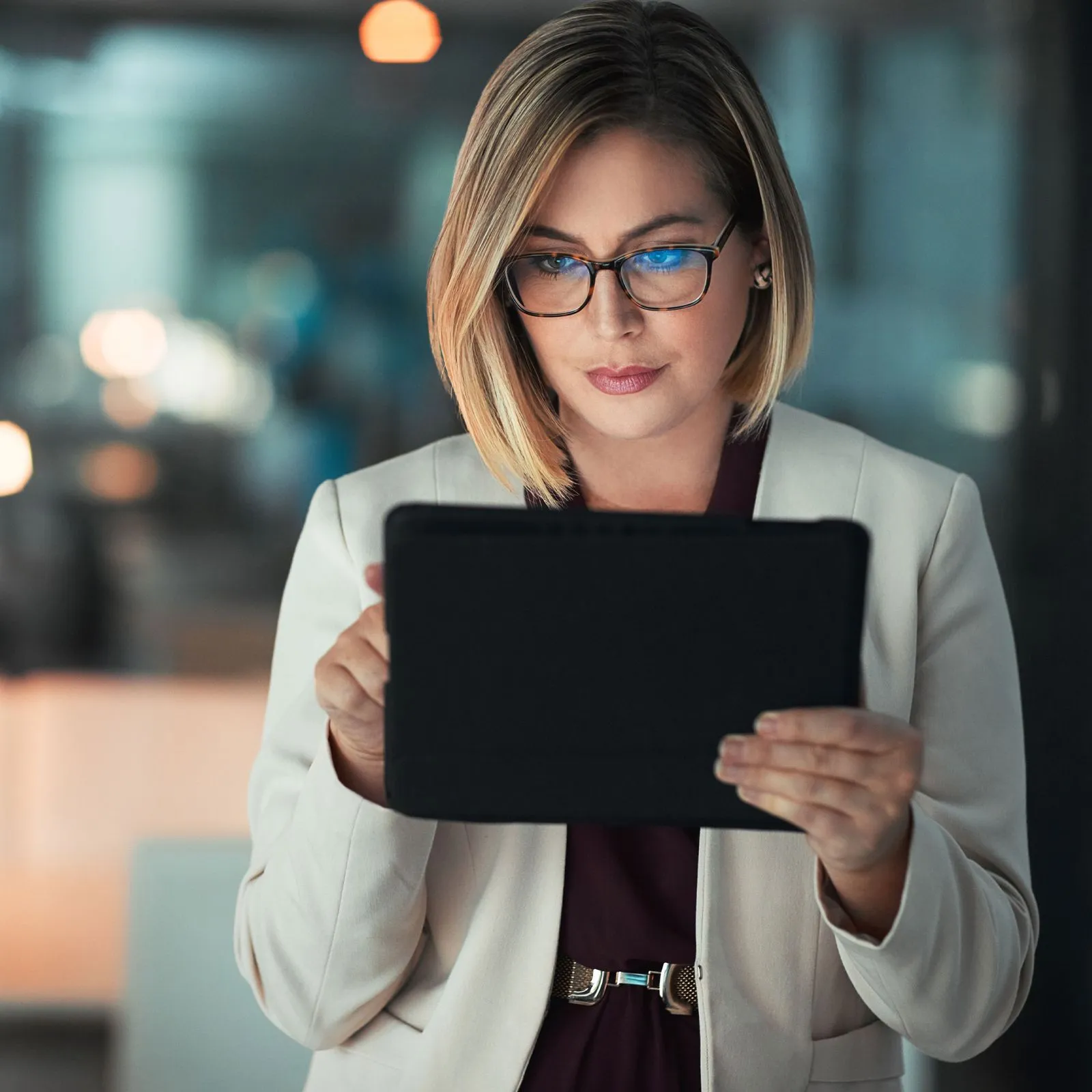 Woman with short blonde hair and glasses uses a tablet in a softly lit office setting.