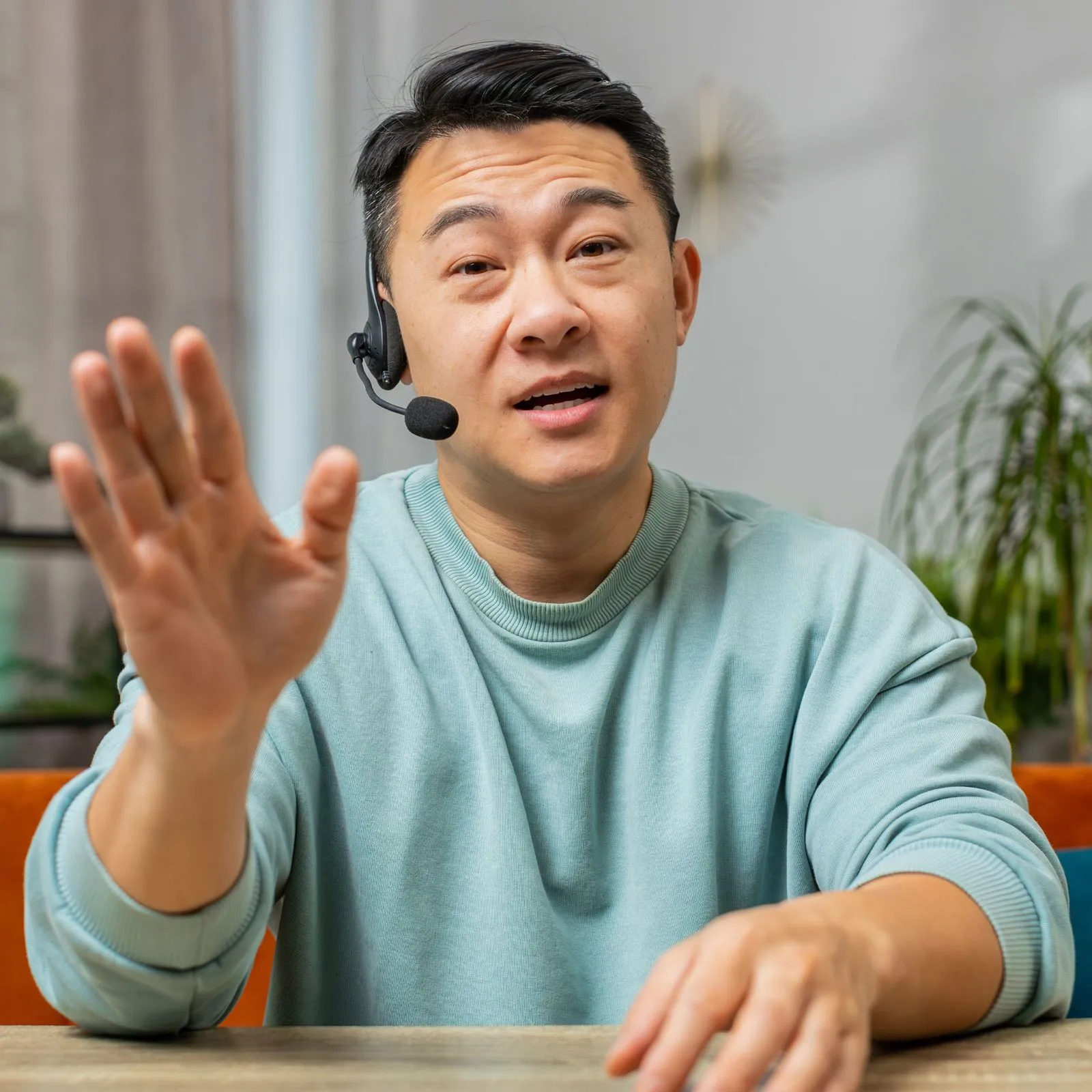Man wearing a headset and speaking with one hand raised in a gesture, seated at a table indoors.