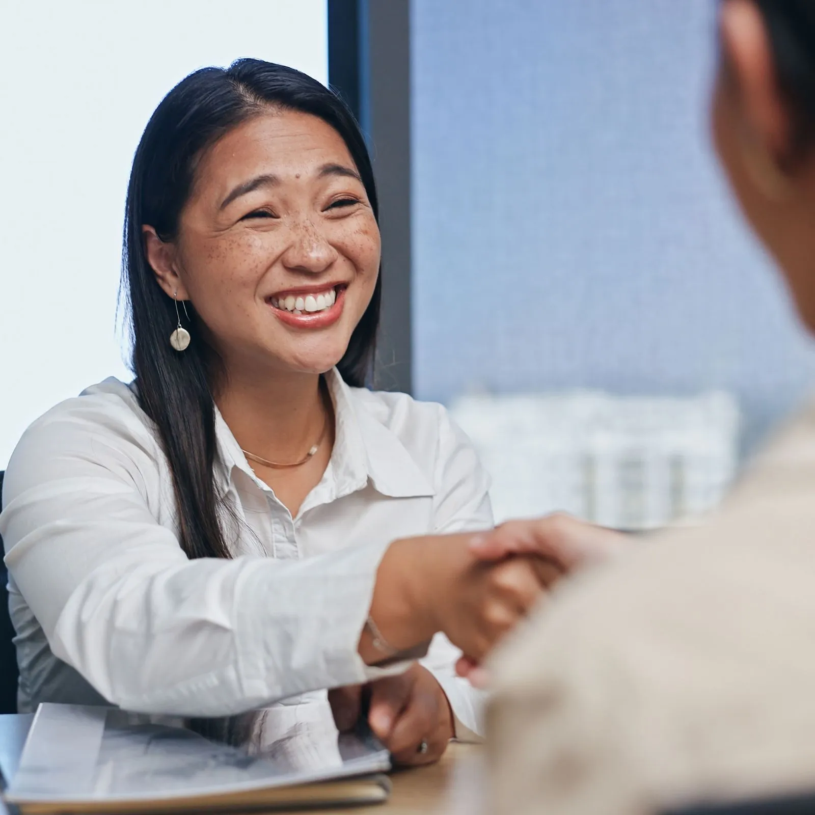 Smiling woman in white shirt shaking hands with another person across a desk.