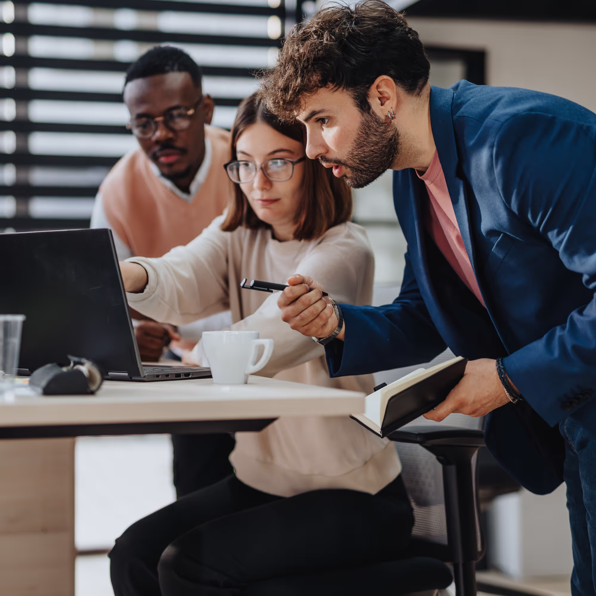 Three coworkers collaborating around a laptop in a modern office, with one pointing at the screen and another holding a notebook.