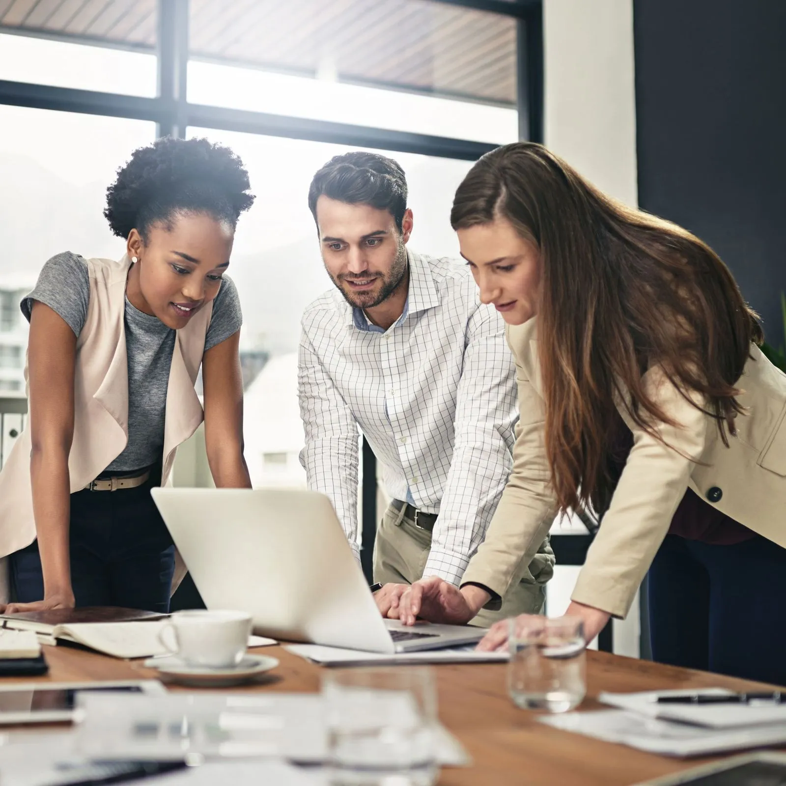 Three colleagues gathered around a table, looking at a laptop screen in a modern office.