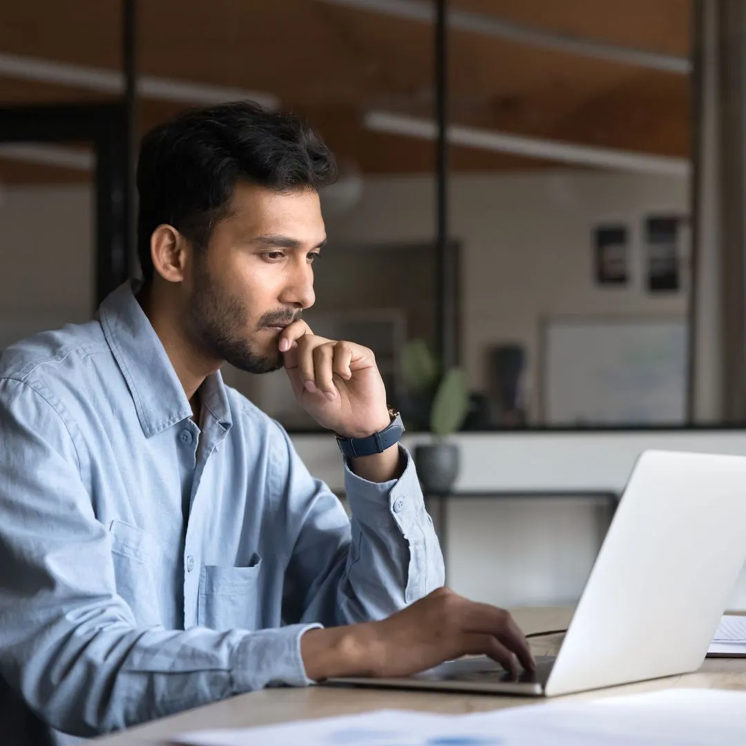Man in light blue shirt focused on working on a laptop at a wooden desk in an office.