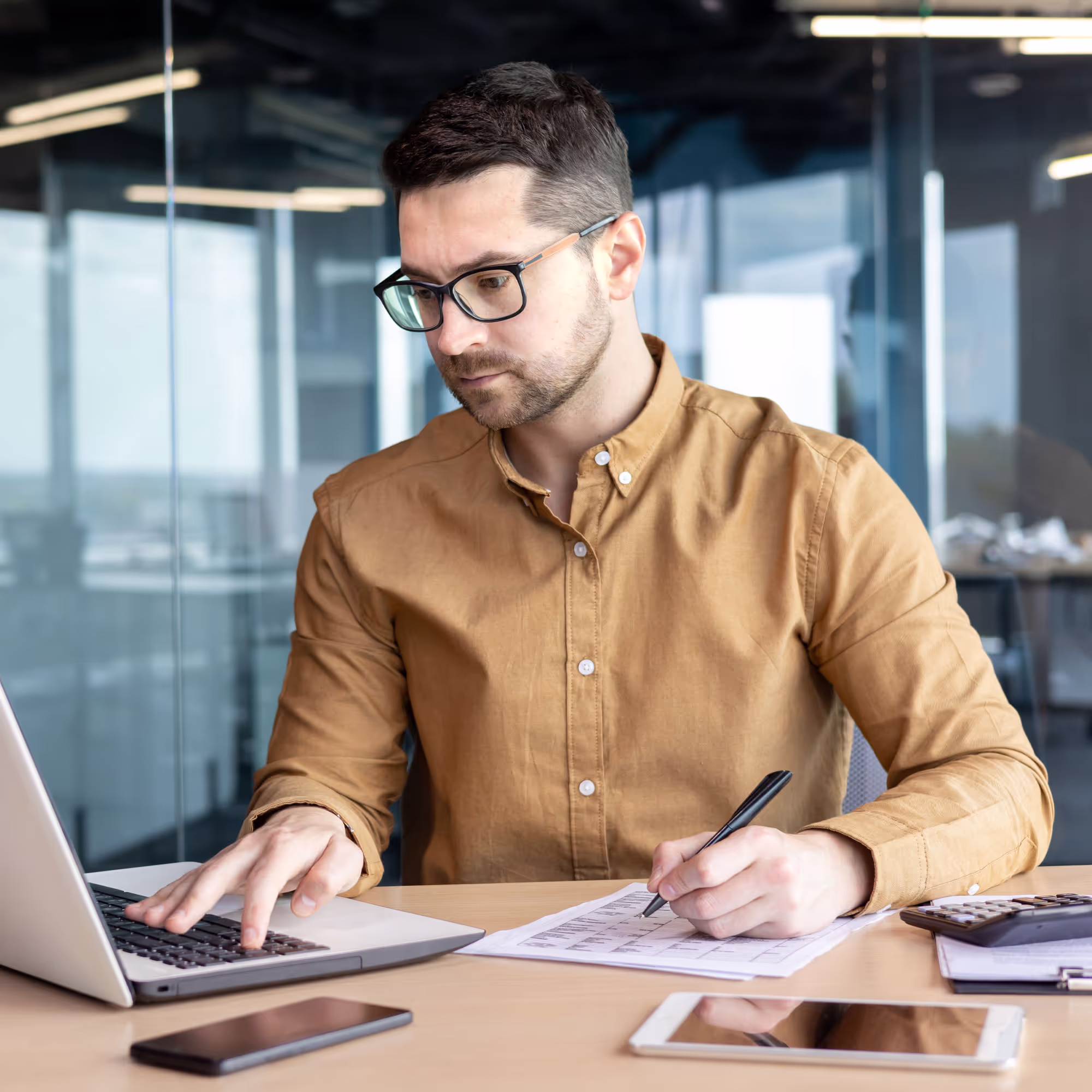 Man wearing glasses and a brown shirt working at a desk with a laptop, writing on paper, with a smartphone and tablet nearby.