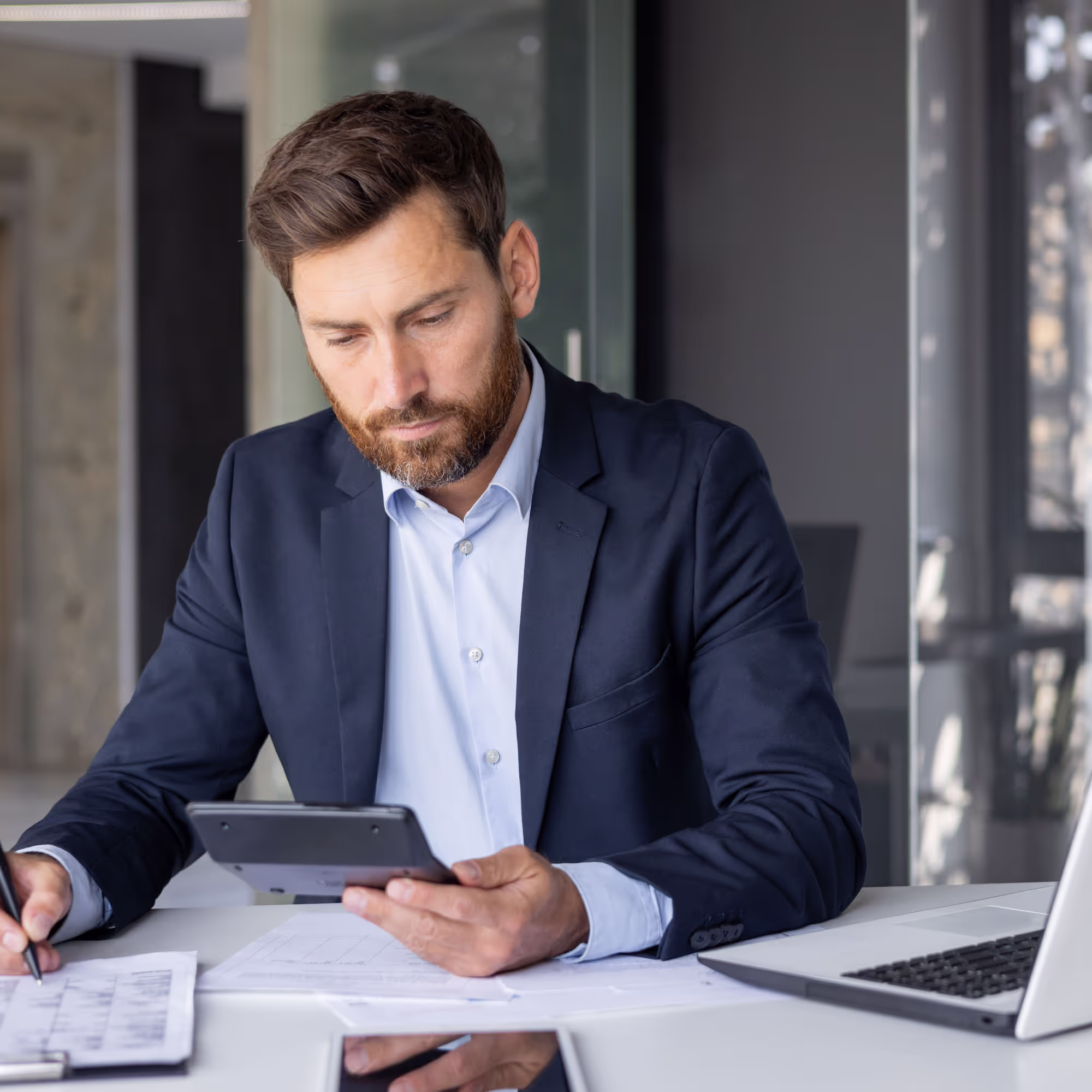 Bearded man in a suit using a calculator and writing on documents at a desk with a laptop.