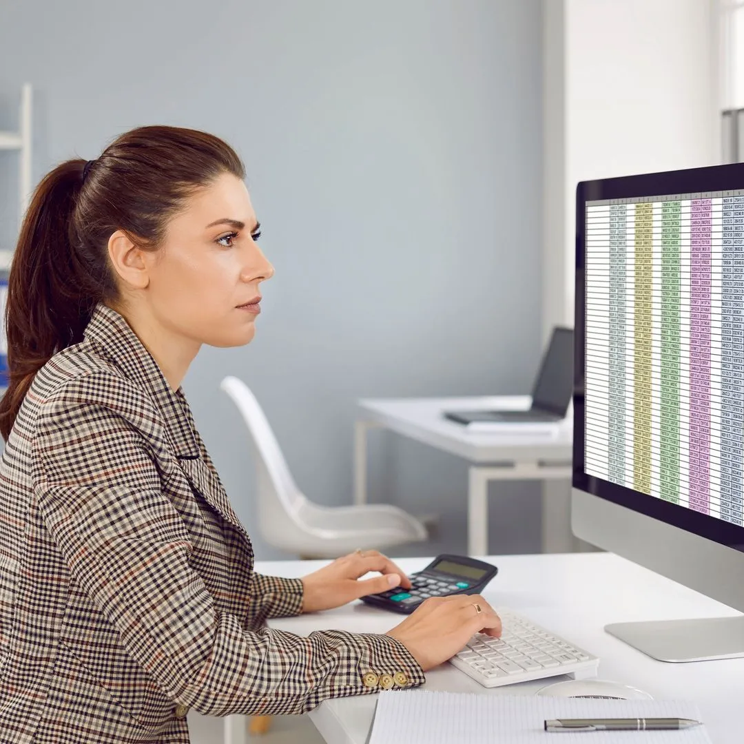 Woman in plaid blazer working at desk with calculator and computer displaying colorful spreadsheet.