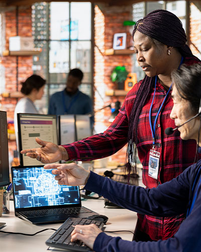 Two women collaborating in an office, one pointing at data visualization on a laptop screen and the other using a keyboard and headset.