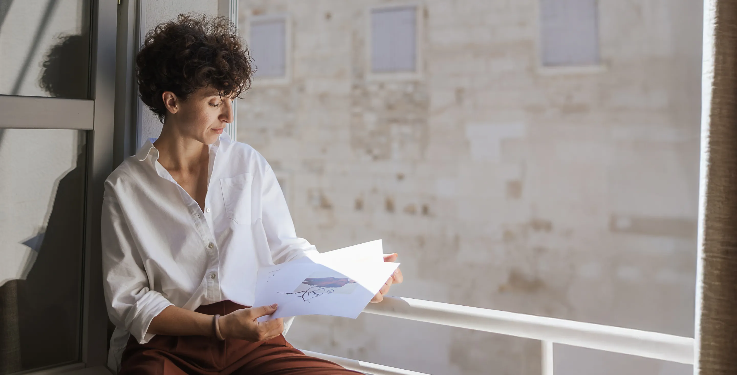 Person with curly hair in a white shirt sitting by a window reading a paper with an illustration.