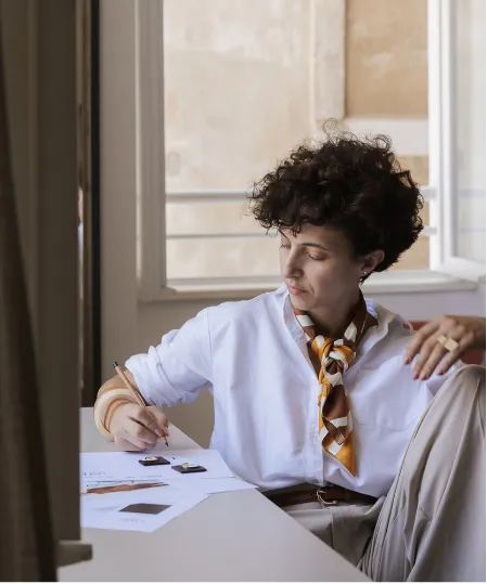 Person with short curly hair and a patterned scarf around their neck sitting at a desk, writing on papers with swatch samples.