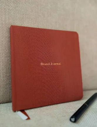 Red hardcover Brand Journal notebook with a ribbon bookmark resting on a beige fabric surface next to a black pen.