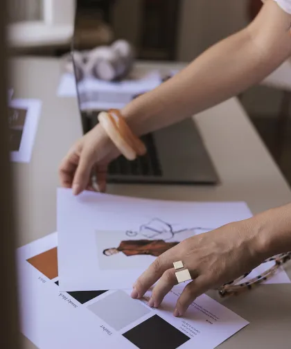 Person arranging fashion design sketches and color swatches on a desk with a laptop in the background.