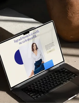 Laptop displaying a website with a photo of a woman in white shirt and blue pants, text in Serbian about building a business led by the buyer, placed on a shadowed surface next to a large pottery vase.