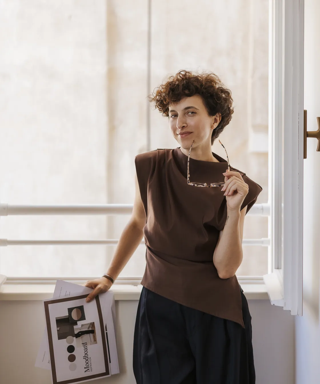 Person with short curly hair holding eyeglasses and a mood board paper, standing by a window with white railings.