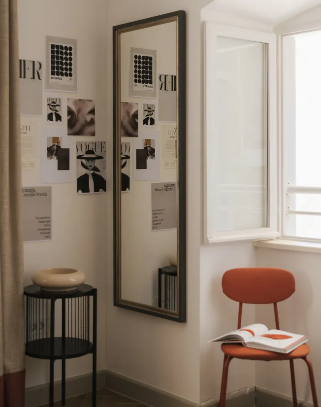Corner of a room with a tall mirror reflecting wall art and a black side table holding a beige ceramic bowl, next to an orange chair with an open book under a window.