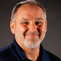 Smiling middle-aged man with short gray hair, beard, and navy shirt against a dark background.