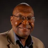 Smiling man wearing glasses and a tan jacket against a dark background.