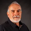 Middle-aged man with gray hair, beard, and mustache wearing a dark shirt against a dark background.